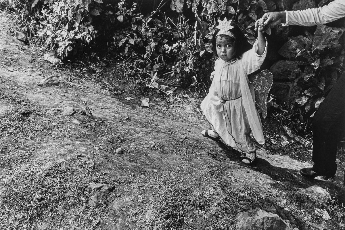 Black and white photo of a child in a dress with wings being held by an adult outdoors.