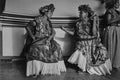 Two women in traditional attire with floral headpieces and skirts, sitting on a bench.
