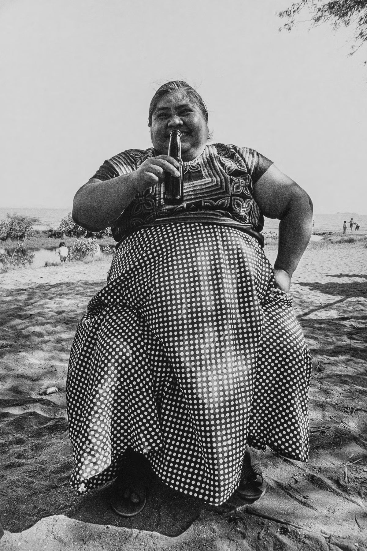 Person in traditional attire sitting outdoors holding a bottle.