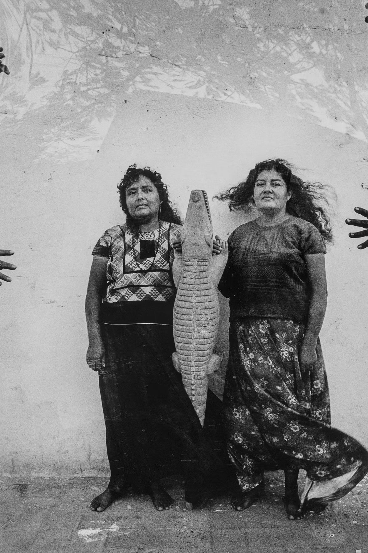 Two women standing side by side with a crocodile between them, against a textured wall.