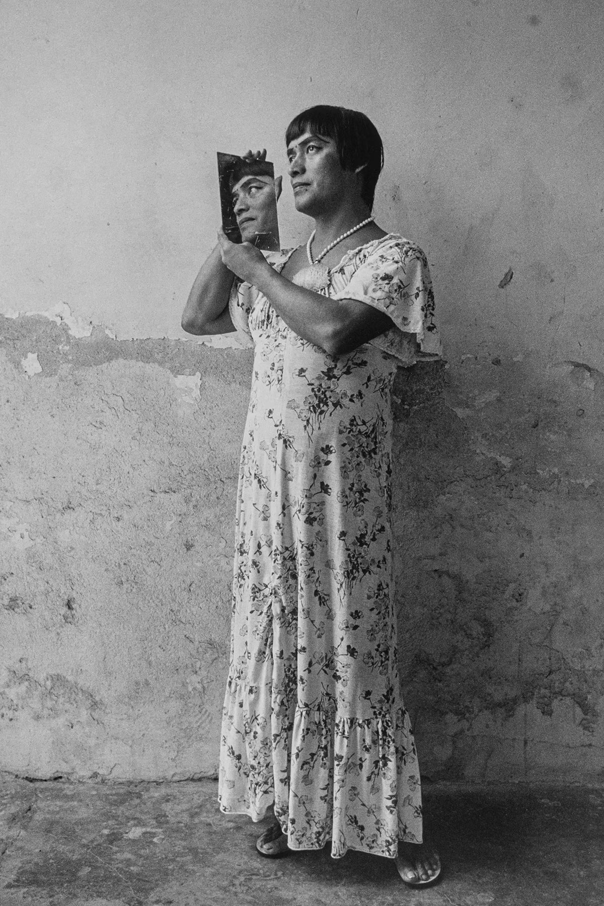 Woman in a floral dress holding a mirror, standing against a textured wall.