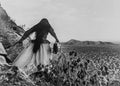 Person in a long dress and headscarf standing on rocky terrain with cacti and desert landscape in the background.