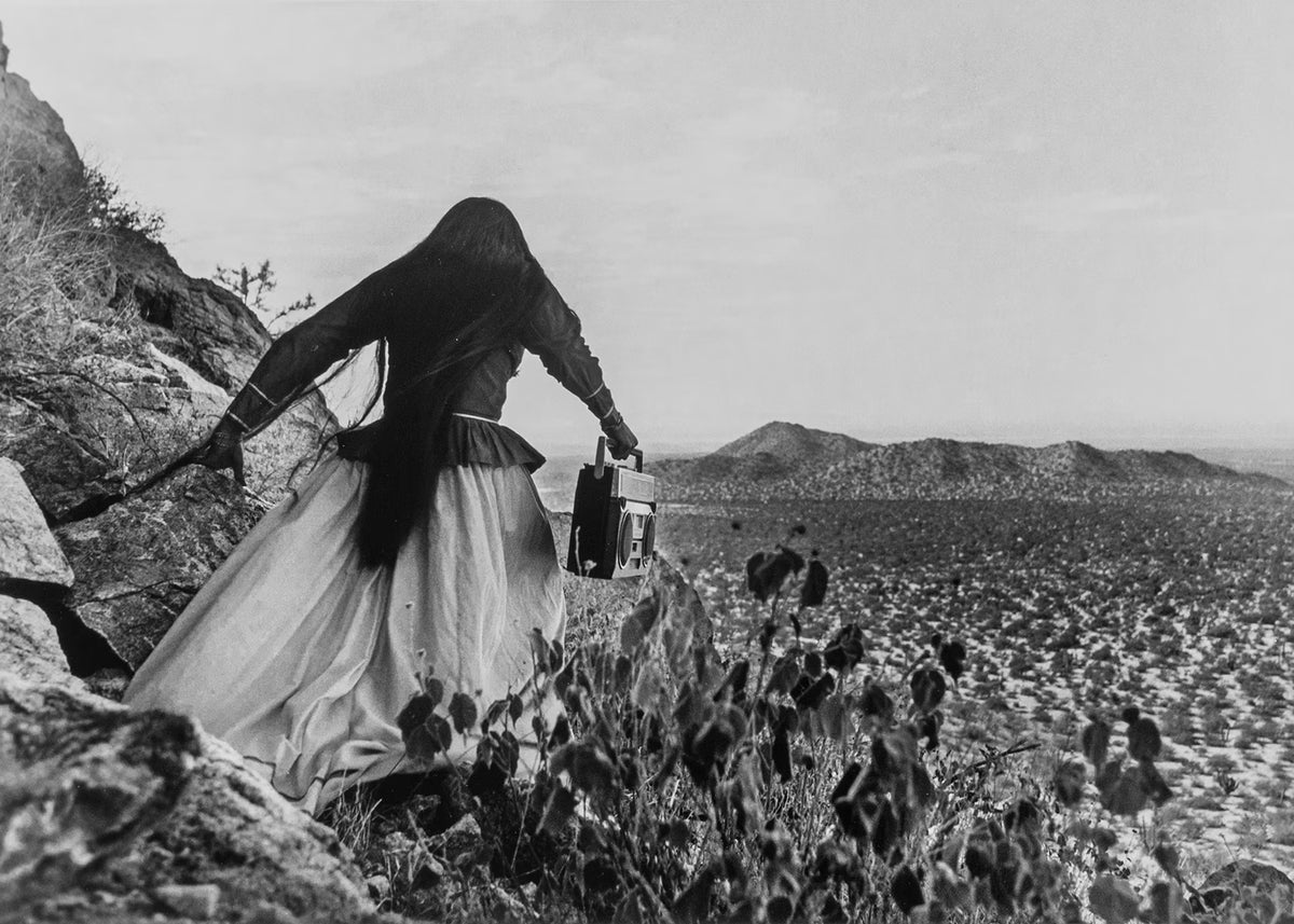 Person in a long dress and headscarf standing on rocky terrain with cacti and desert landscape in the background.