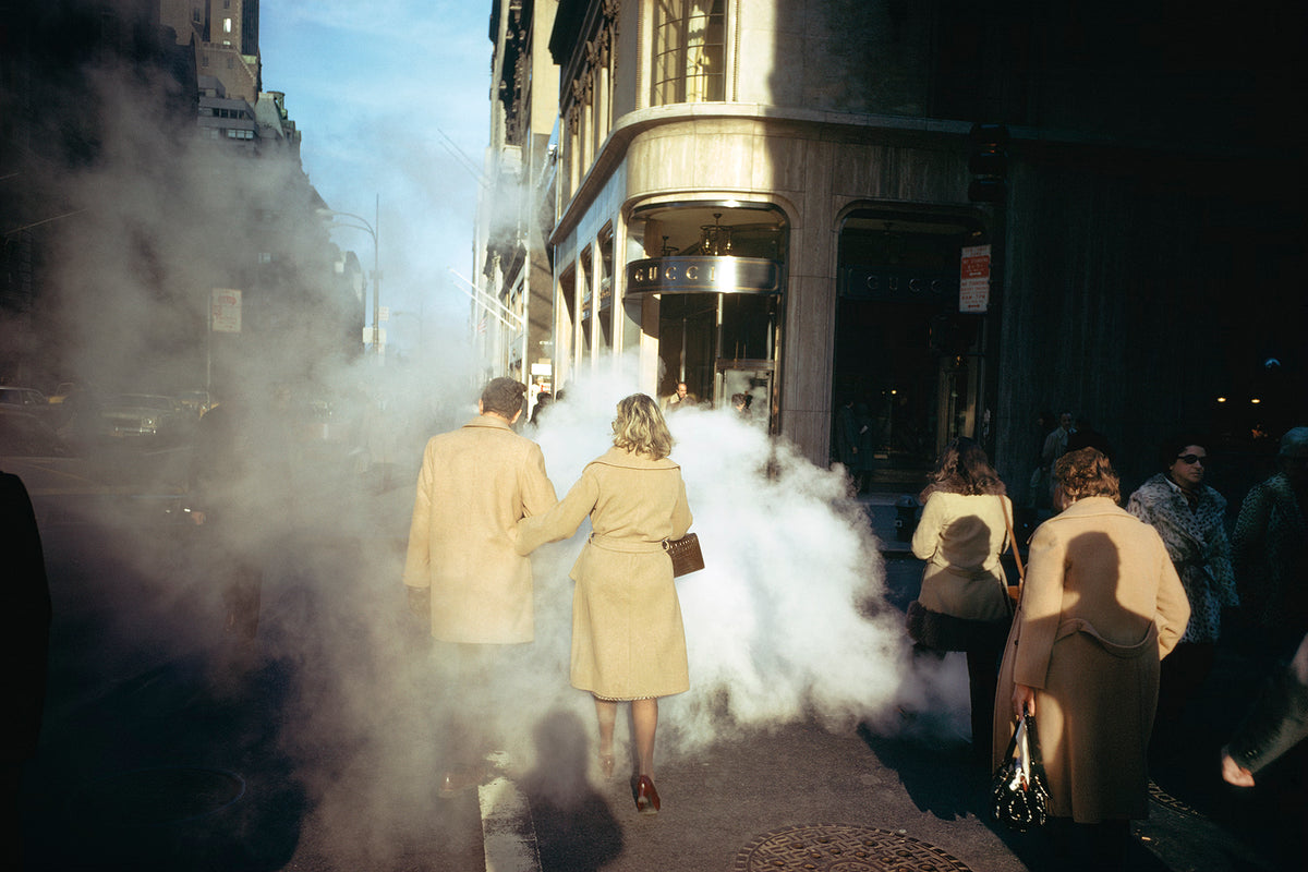 People walking on a city street with steam or smoke rising from a source.
