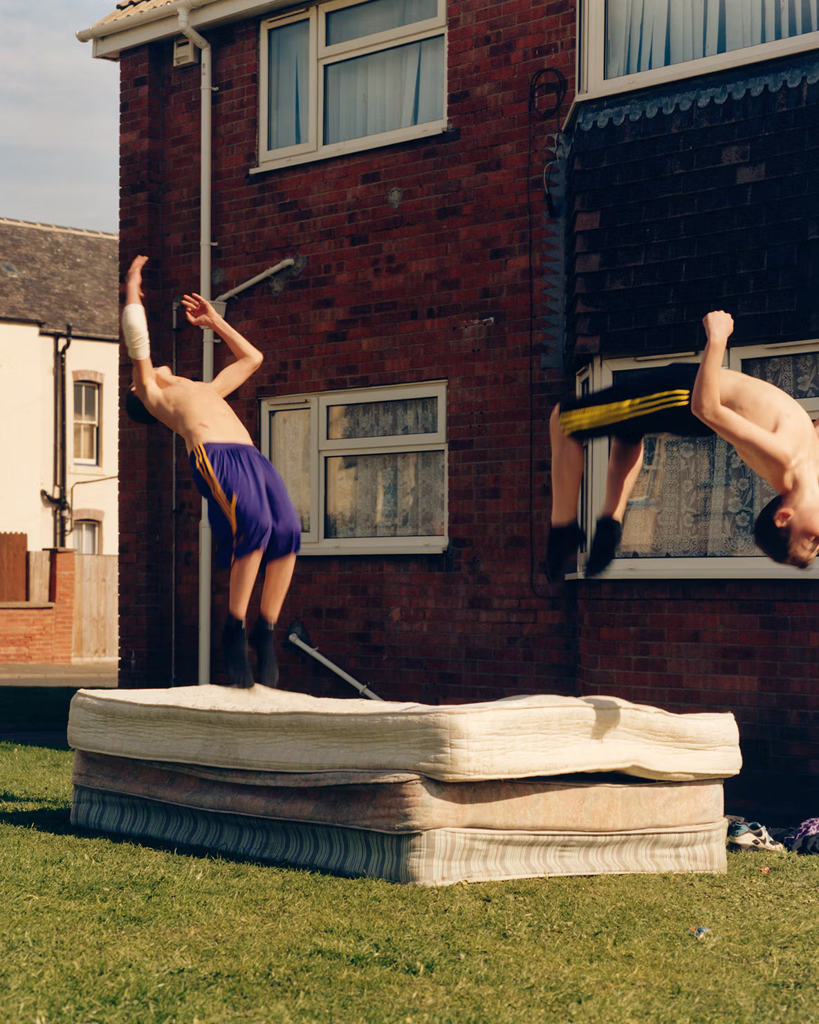 Two young men performing acrobatics on a mattress in front of a brick building.